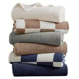 A stack of folded towels in various colors and patterns, including solid white, beige, gray, and blue, with some featuring checkered designs.