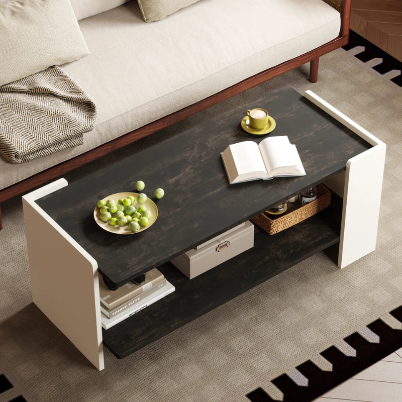 A modern coffee table with a black top, featuring a book, a coffee cup, and a plate of green fruits. Below are two shelves with boxes and a woven basket, positioned in front of a beige sofa with cushions.