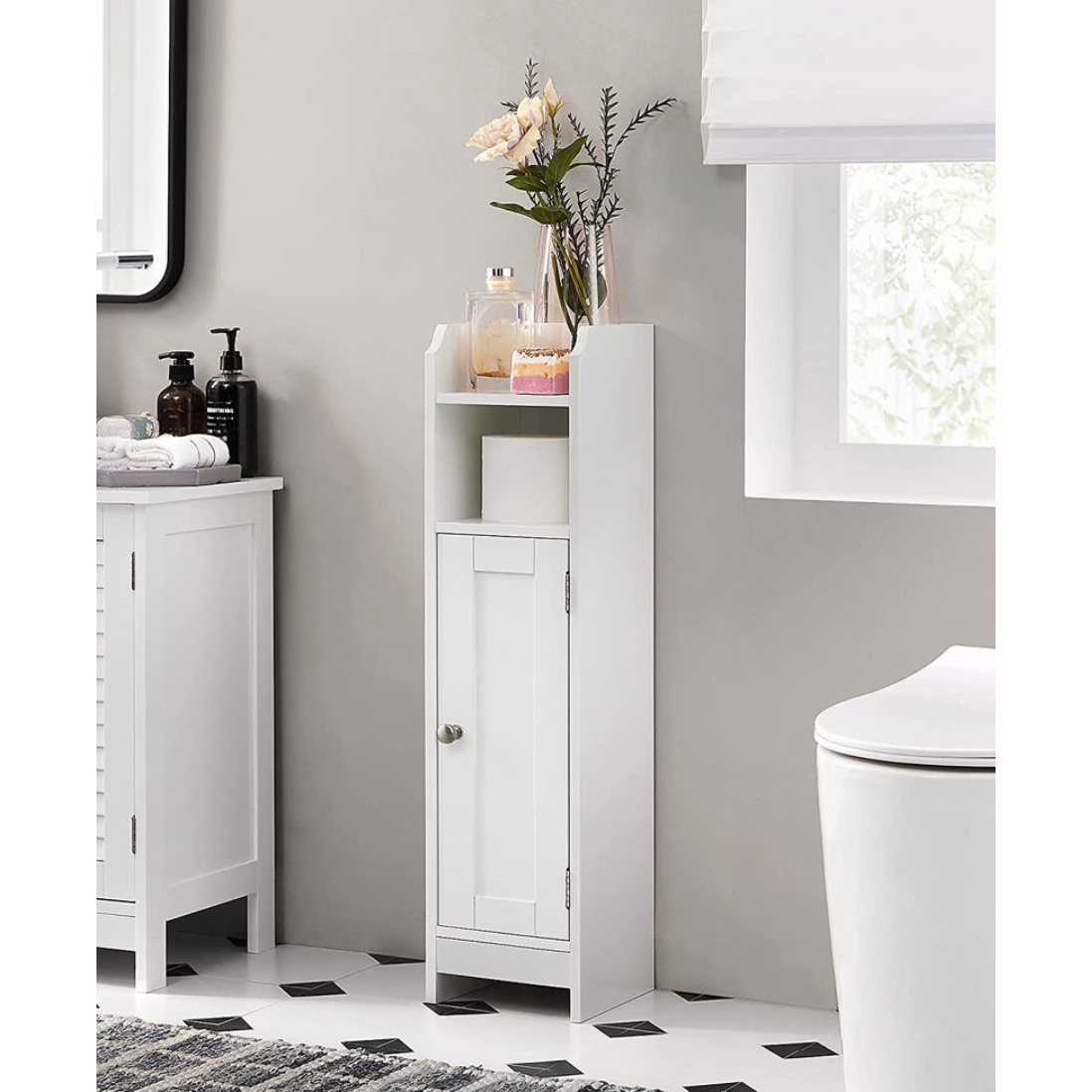 A slim white bathroom cabinet with a small door at the bottom and an open shelf above, holding a roll of toilet paper and decorative items like a vase with flowers and a candle. It's positioned on a patterned black and white floor next to a sink.