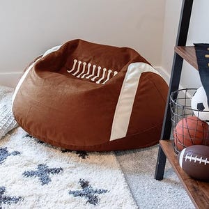 A bean bag chair styled like an American football with brown fabric, white laces, and stripes. It sits on a patterned rug in a corner near sports-themed decor.