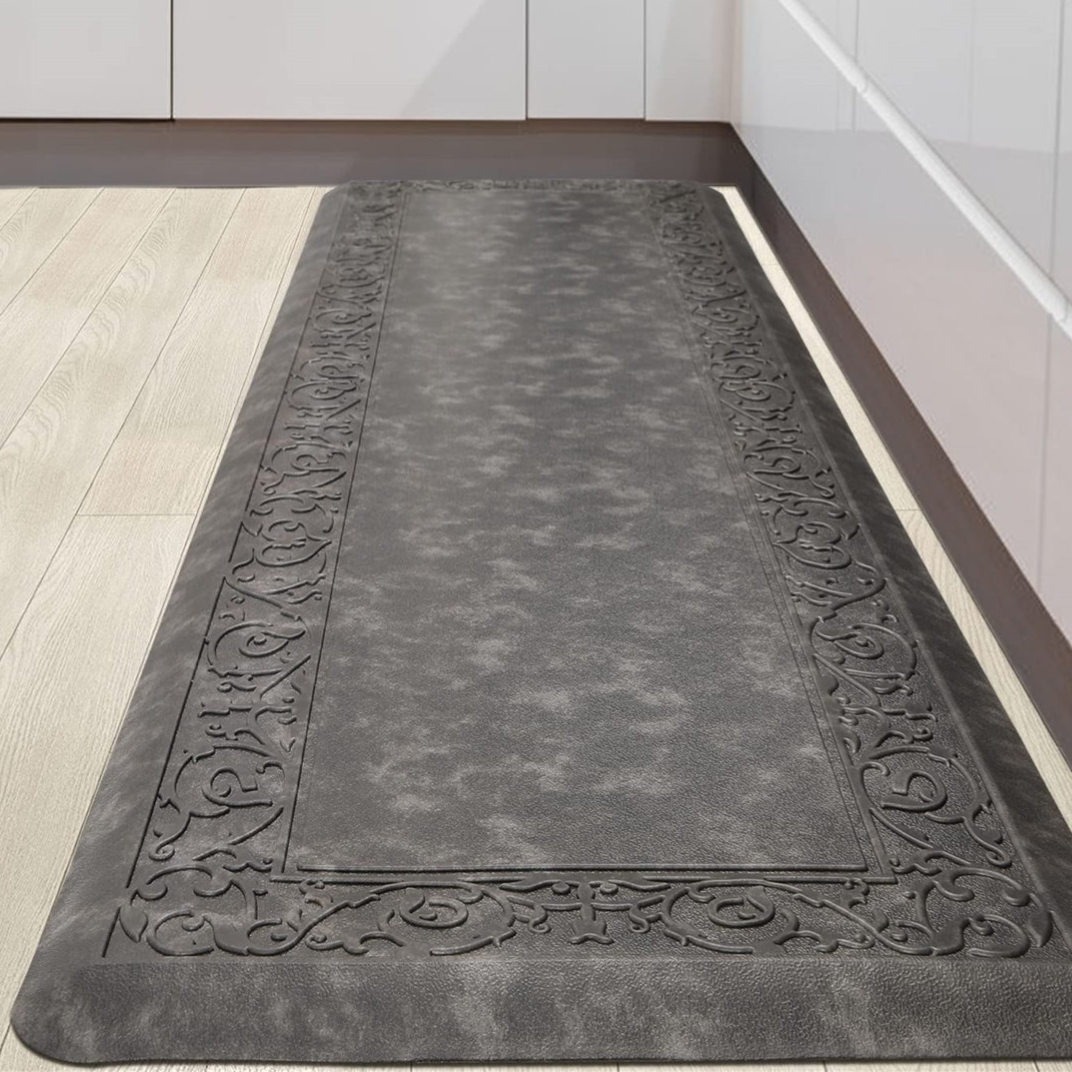 A dark gray anti-fatigue kitchen mat with an ornate border design, placed on a light wood floor in front of white cabinets.