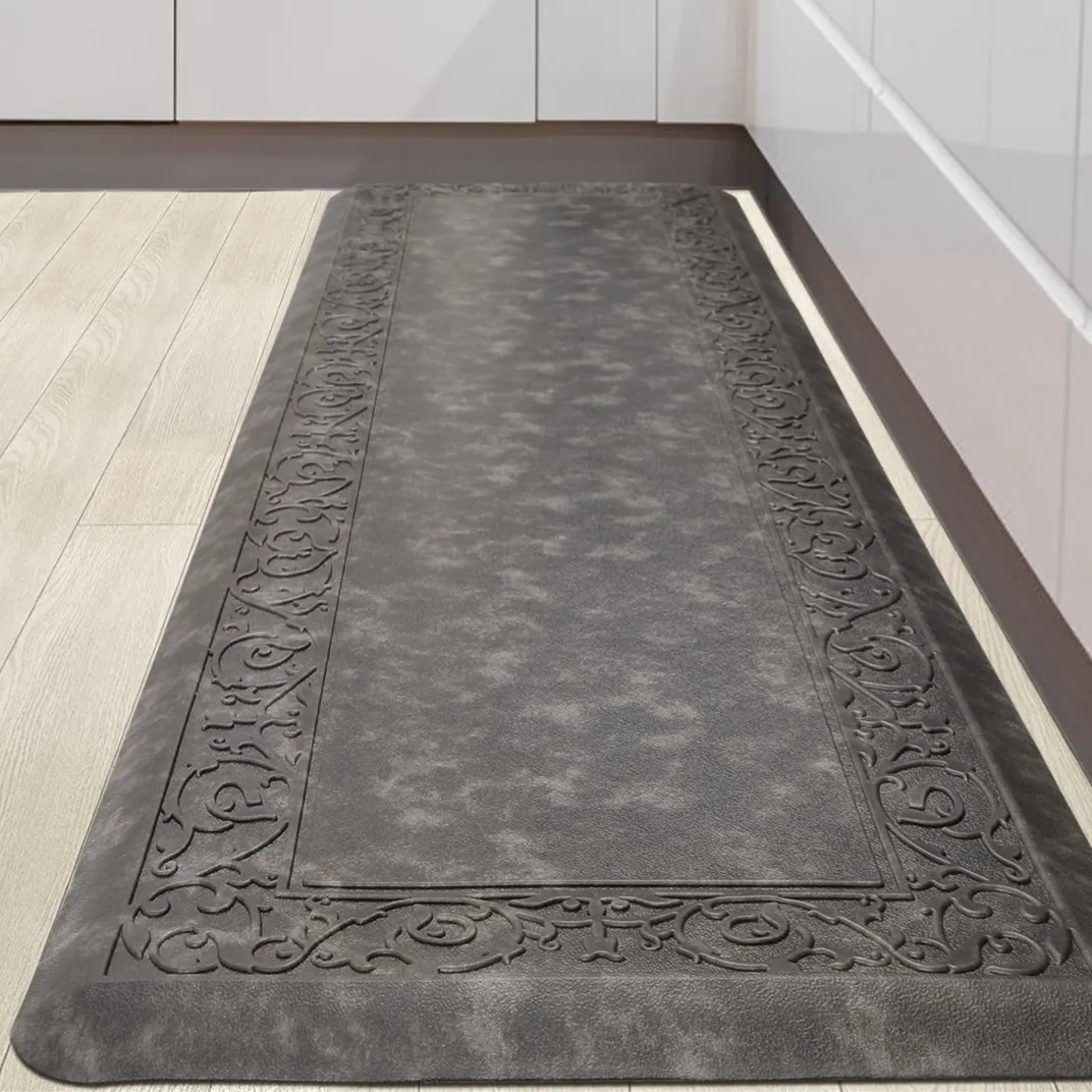 A dark gray anti-fatigue kitchen mat with an ornate border design, placed on a light wood floor in front of white cabinets.