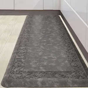 A dark gray anti-fatigue kitchen mat with an ornate border design, placed on a light wood floor in front of white cabinets.