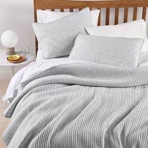 A light gray ribbed bedspread and matching pillow shams are set on a wooden bed frame, accompanied by a side table with a lamp, glass of water, and a bowl of cut fruit.