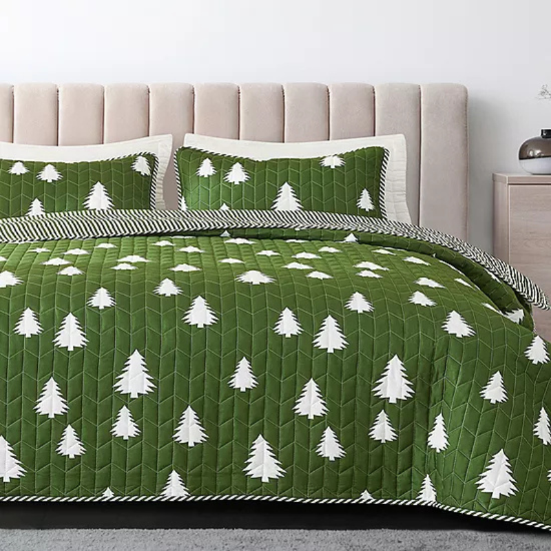 A green quilted bedding set with white tree patterns, featuring a bedspread and two matching pillow shams on a light pink upholstered headboard.