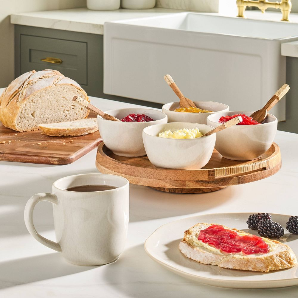 A breakfast setting with a slice of bread spread with red jam, a mug of coffee, a sliced loaf on a cutting board, and a turntable holding four bowls with various spreads including jam and butter. Blackberries garnish the plate.