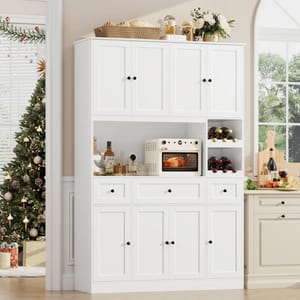 White kitchen pantry featuring multiple cabinets, drawers, a microwave shelf, and wine storage. Decor includes a basket, jars, wine bottles, and floral arrangements. A Christmas tree and gifts are in the background, with a window view of greenery.