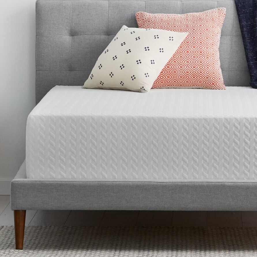 A grey upholstered bed frame with a textured white sheet and two decorative pillows, one cream with blue patterns and one red with geometric designs.