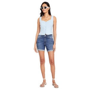 A woman is wearing a light blue sleeveless buttoned top with dark denim shorts, white strappy sandals, and white sunglasses.