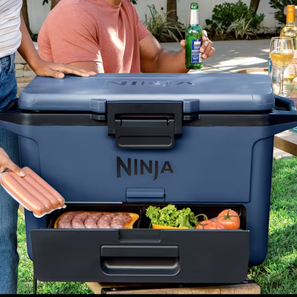 A Ninja cooler with storage features an open drawer displaying sausages, lettuce, and tomatoes, with a person holding hot dogs and someone else holding a beverage.