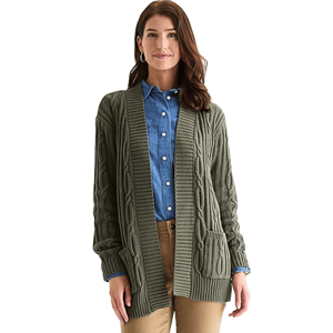 A woman is wearing an olive green cable knit cardigan over a blue denim button-up shirt and beige pants. The cardigan has two front pockets and a ribbed hem.