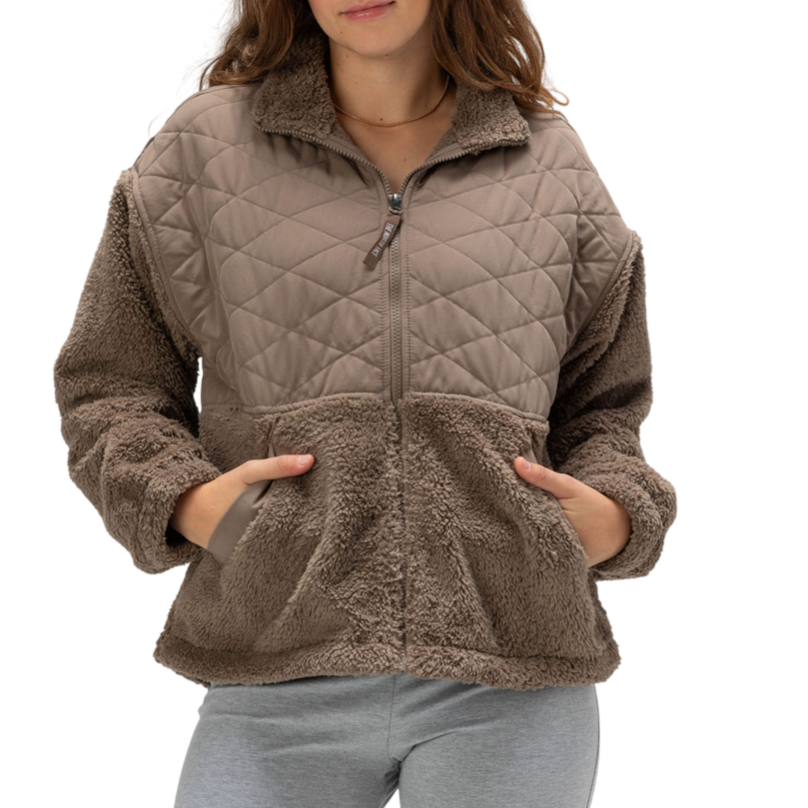 A woman is wearing a taupe, quilted, and faux-shearling jacket with a zipper front and pockets, paired with light gray leggings.