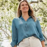A person is wearing a light blue denim shirt and cream-colored pants, standing outdoors with trees in the background.