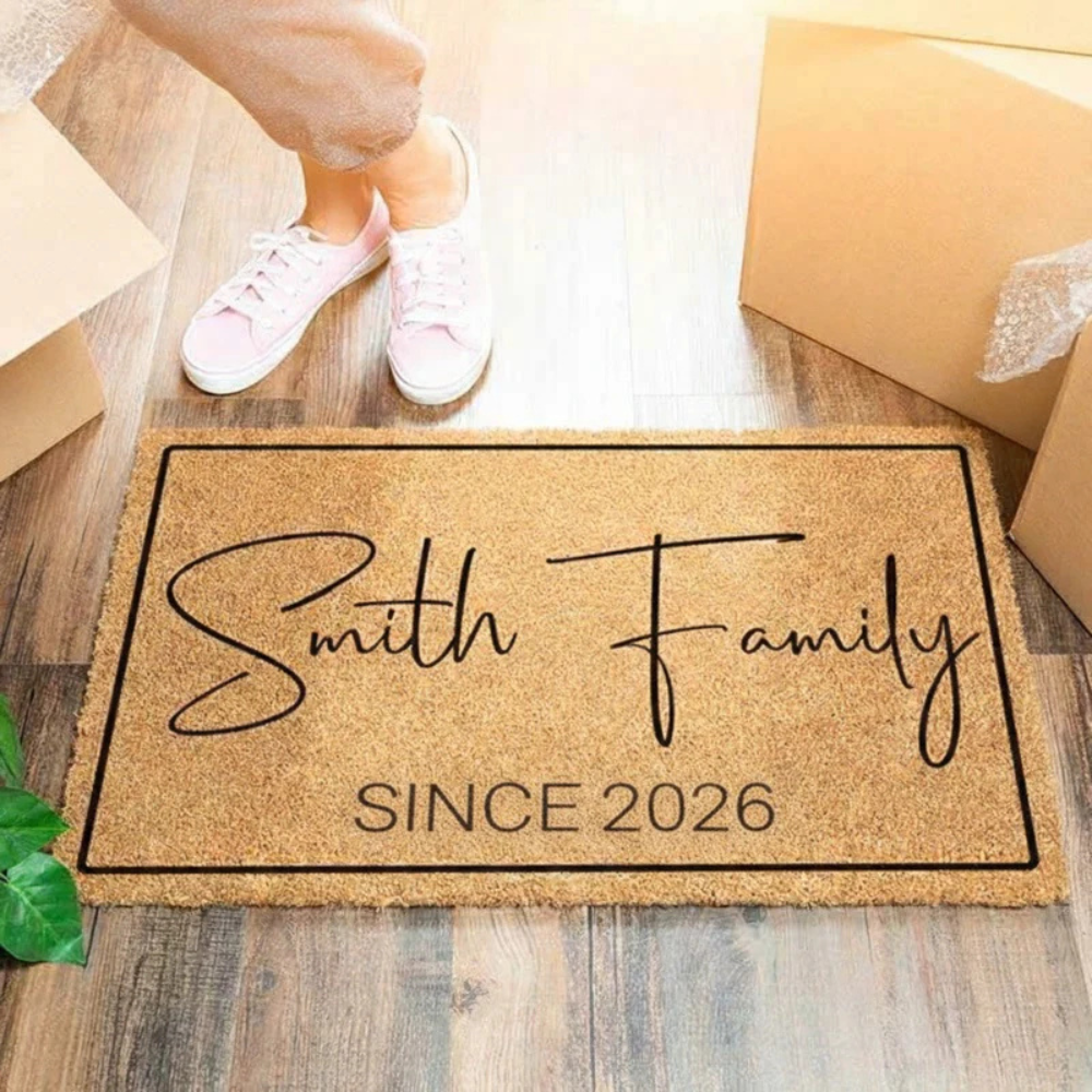 A doormat with the text \“Smith Family Since 2026\“ is on a wooden floor surrounded by unpacked boxes and legs in pink sneakers standing nearby.