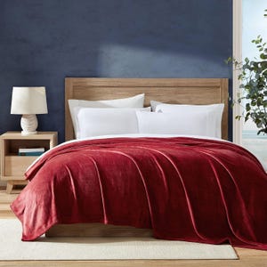 A red velvet comforter on a bed with white sheets, wooden headboard, beside a lamp on a nightstand.