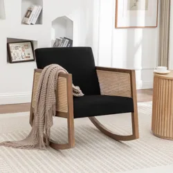A black cushioned rocking chair with wooden armrests and woven rattan sides, styled with a taupe throw blanket, is set on a light patterned rug next to a round wooden side table holding a coffee cup.
