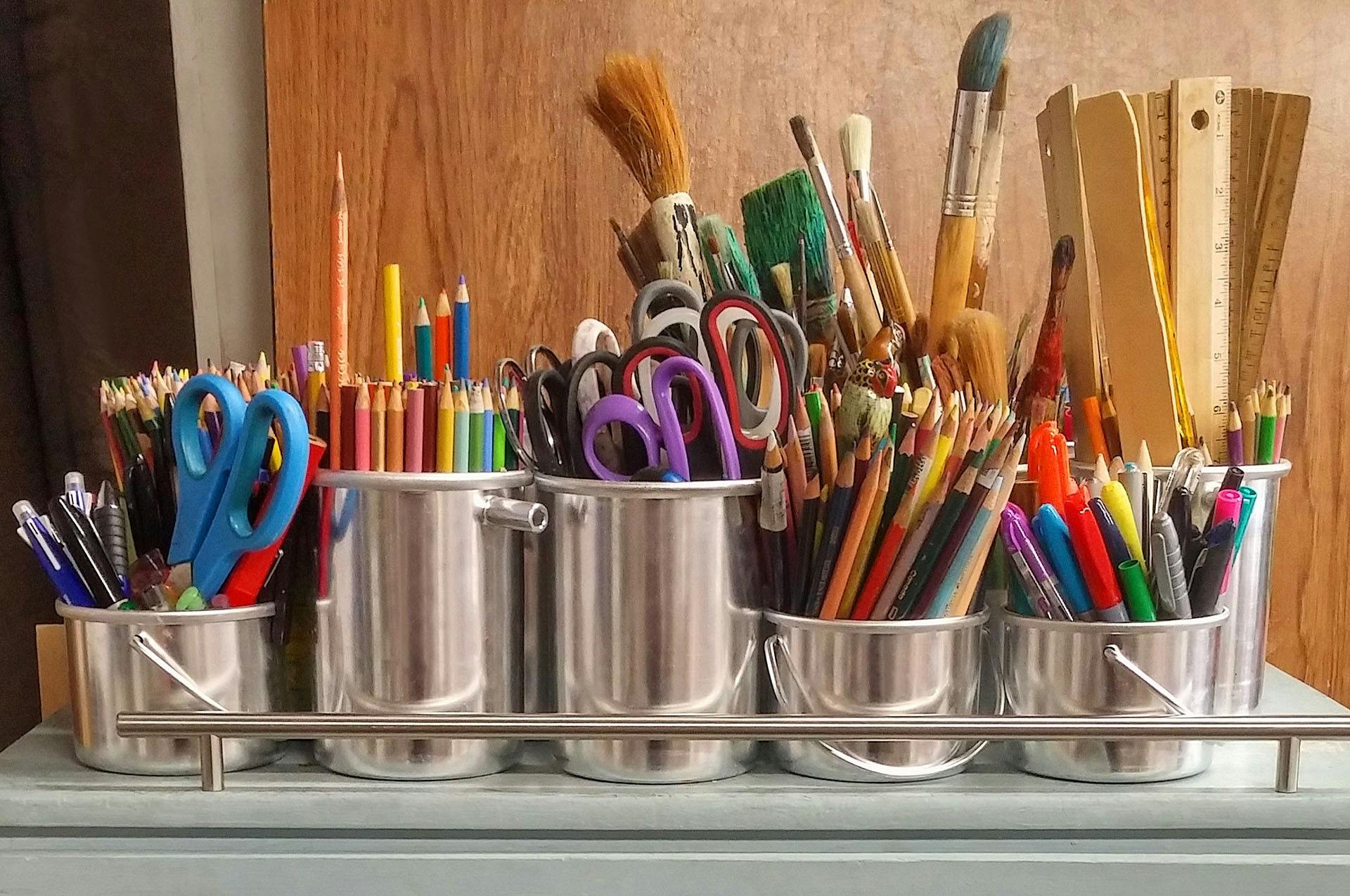 A variety of art supplies, including colored pencils, paintbrushes, scissors, and rulers in metal containers.