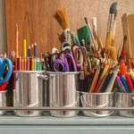 A variety of art supplies, including colored pencils, paintbrushes, scissors, and rulers in metal containers.