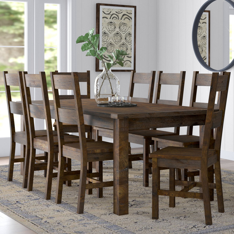 A wooden dining table set with six chairs, featuring a rustic design. Decor includes a glass vase with green leaves and patterned artworks on the walls.