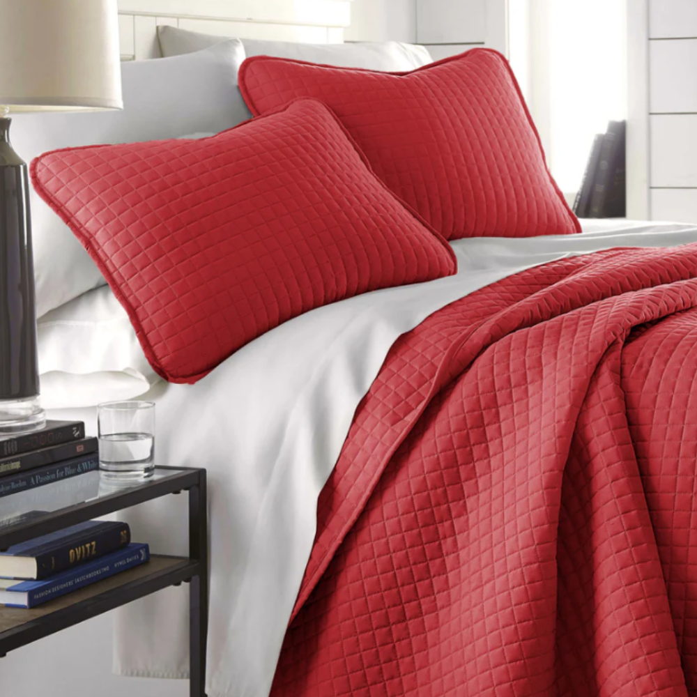 Red quilted pillow shams and a matching quilt cover a bed with white sheets, next to a black side table with books and a glass of water.