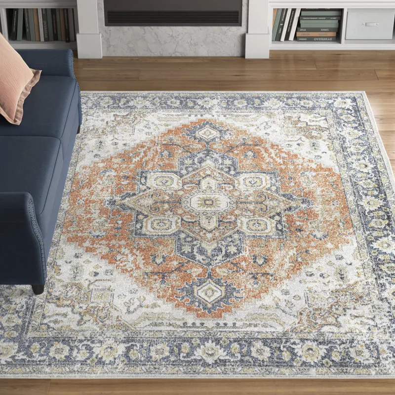 A decorative rug with a geometric and floral design in shades of blue, orange, and white, placed on a wooden floor next to a blue sofa with a pink pillow.