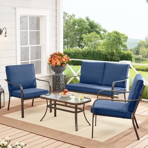 Outdoor patio furniture set with two blue cushioned chairs, a blue cushioned loveseat, and a glass-top coffee table, set on a deck. The arrangement includes decorative flowers and drinks, creating a cozy outdoor seating area.