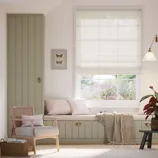 A cozy room with a cushioned window seat, a built-in wardrobe, a rattan armchair, and a small side table. The decor includes soft neutral tones, a wall-mounted light, and botanical accents. A framed butterfly art piece adds a decorative touch.