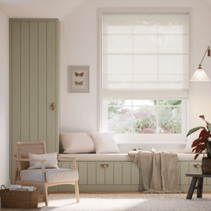 A cozy room with a cushioned window seat, a built-in wardrobe, a rattan armchair, and a small side table. The decor includes soft neutral tones, a wall-mounted light, and botanical accents. A framed butterfly art piece adds a decorative touch.