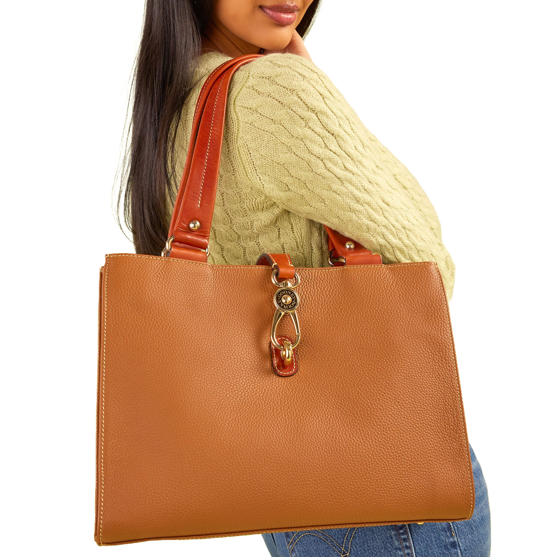A tan leather tote bag with double straps and a gold-tone logo lock detail on the front, featuring reinforced edges and visible stitching. The bag is worn over the shoulder, complementing a cable-knit sweater and blue jeans.