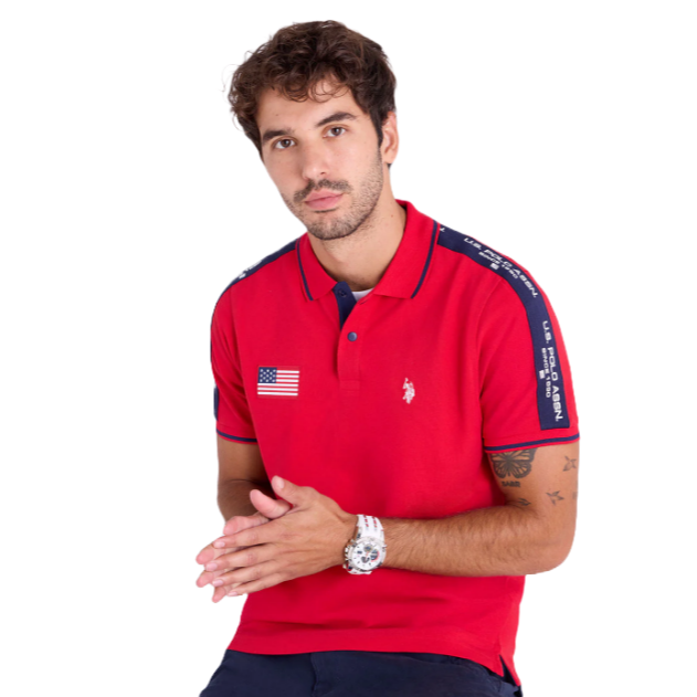 A man wears a red U.S. Polo Assn. polo shirt featuring an American flag patch and branding on the sleeves, paired with dark pants.