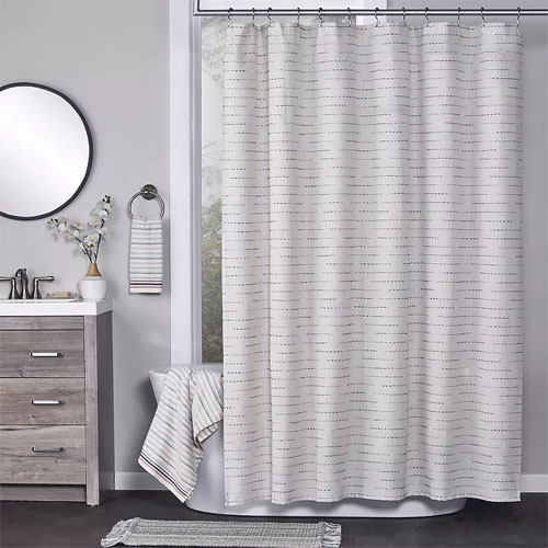 A bathroom features a striped white shower curtain, a round mirror, a wooden vanity with a towel holder, a striped hand towel, a faucet with a soap dispenser, and an orchid decoration. A matching striped towel and textured mat complete the decor.