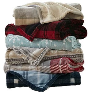 A stack of six folded blankets featuring various patterns, including plaid, snowflakes, and geometric designs, in colors like beige, red, blue, and grey.