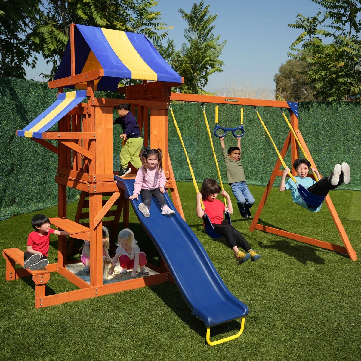 A children's playset features a wooden structure with a blue and yellow canopy, a slide, swings, and a small sandbox area. Several children are playing on the set in a grassy yard.
