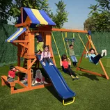 A children's playset features a wooden structure with a blue and yellow canopy, a slide, swings, and a small sandbox area. Several children are playing on the set in a grassy yard.