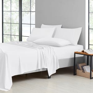 A neatly made bed with white sheets, pillowcases, and a light blanket, next to a bedside table with a glass and eyeglasses.