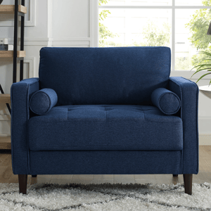 Blue loveseat with cylindrical bolster pillows, tufted seat, and wooden legs, situated on a light-colored, textured rug in a room with bookshelves and large windows.