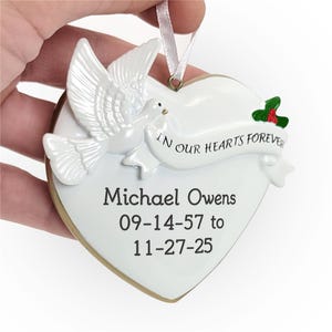 A heart-shaped memorial ornament featuring a white dove, with the inscription \“IN OUR HEARTS FOREVER.\“ Engraved below are the name Michael Owens and dates 09-14-57 to 11-27-25.