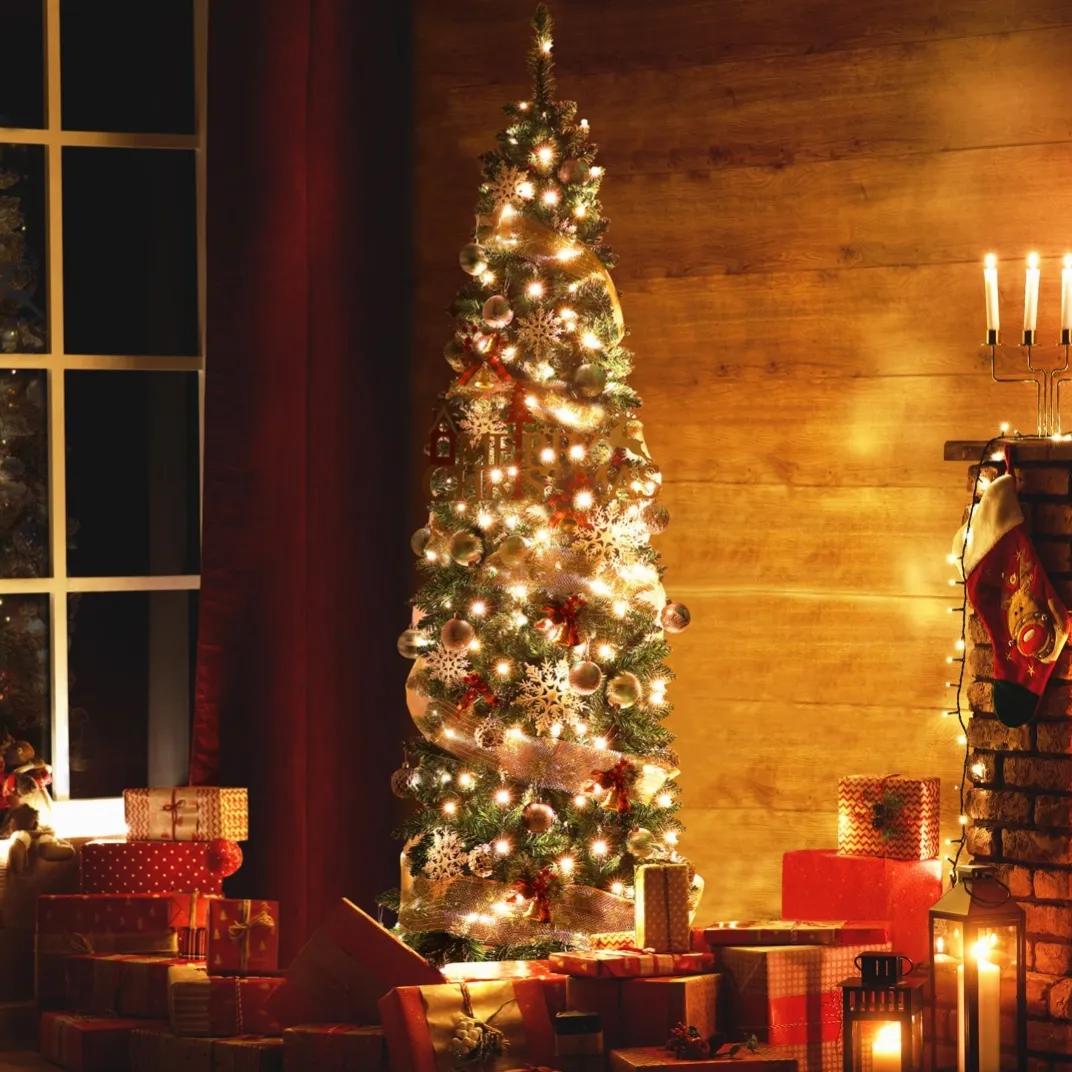A decorated Christmas tree with lights and ornaments stands beside a brick fireplace, surrounded by wrapped presents and candle-lit lanterns. A stocking hangs from the mantel, enhancing the cozy, festive atmosphere.
