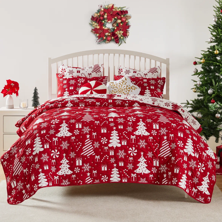 A festive bedding set with a red Christmas-themed pattern featuring trees, presents, and snowflakes. The room is decorated with a wreath, a Christmas tree, and poinsettias, creating a holiday ambiance.