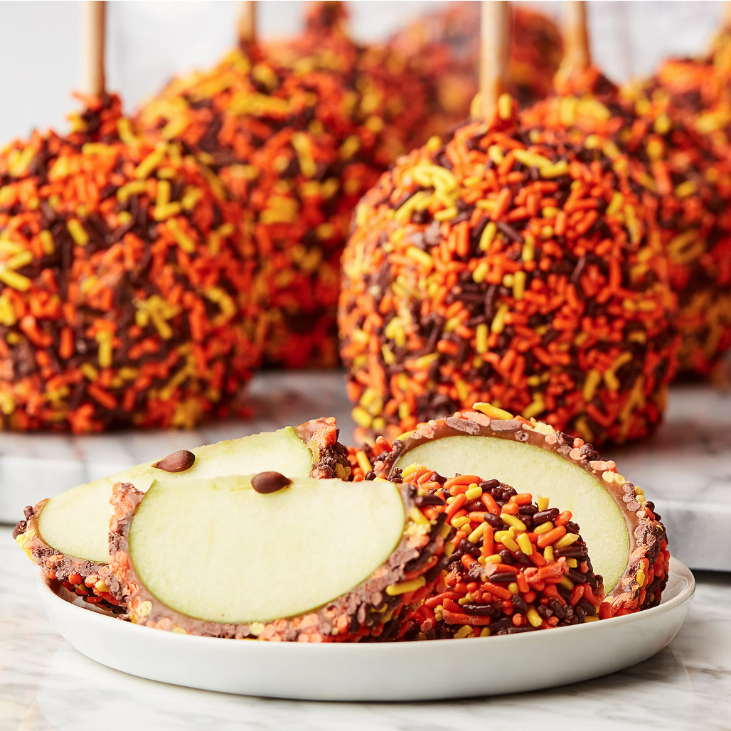 Caramel apples coated with orange, yellow, and brown sprinkles are displayed, with sliced pieces on a plate.