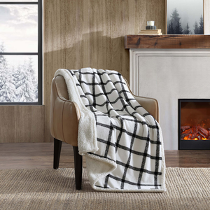 A brown chair draped with a black-and-white checkered fleece and sherpa throw blanket sits in a cozy room with a fireplace, wooden walls, and a snowy view outside.