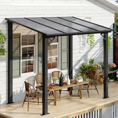 A metal patio pergola with a clear roof shelters a wooden dining table and chairs on a deck, surrounded by plants and flowers against a white house exterior with green shuttered windows.