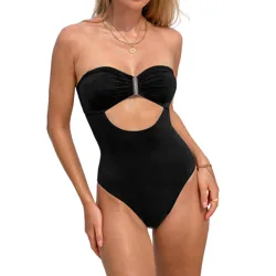A blonde woman is wearing a black, strapless one-piece swimsuit featuring a cutout below the bust with a metal accent at the center. She accessorizes with gold hoop earrings and layered necklaces, standing against a beach backdrop.