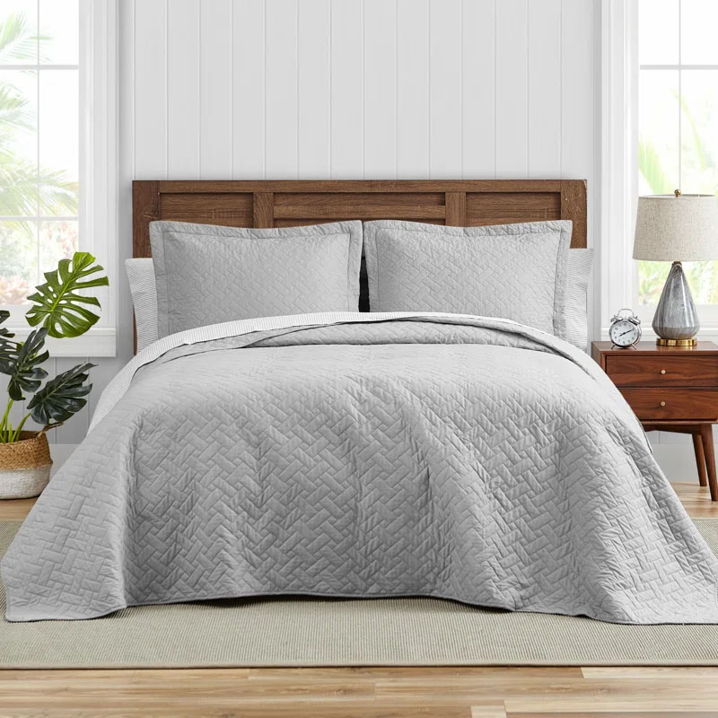 The Tommy Bahama Raffia Cotton Quilt Set includes a light gray quilt and matching pillow shams with a subtle herringbone pattern. It's displayed on a bed with a wooden headboard, side table, lamp, clock, and decorative plant, creating a cozy bedroom setting.