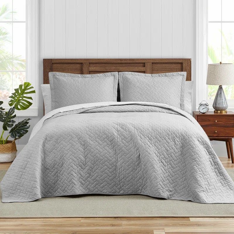 The Tommy Bahama Raffia Cotton Quilt Set includes a light gray quilt and matching pillow shams with a subtle herringbone pattern. It's displayed on a bed with a wooden headboard, side table, lamp, clock, and decorative plant, creating a cozy bedroom setting.