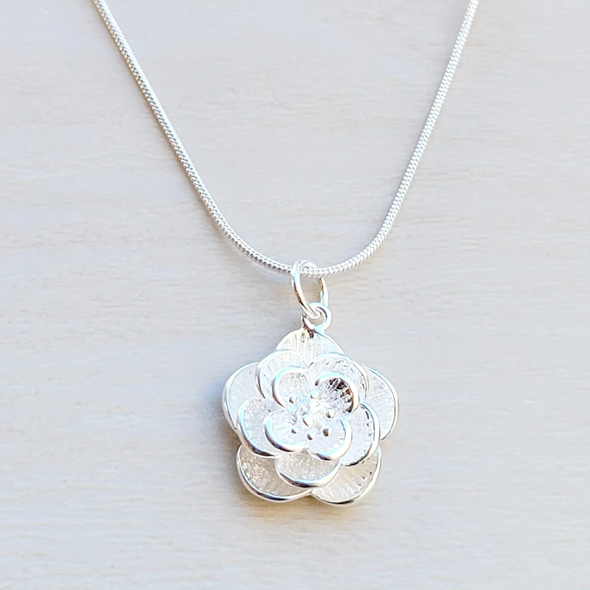A silver necklace features a pendant designed as a layered flower with textured petals, attached to a sleek chain.