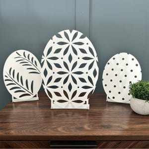 Three white tabletop decorations in the shape of Easter eggs feature intricate cut-out designs: a leaf pattern, a floral pattern, and polka dots, all set on small stands on a wooden table.
