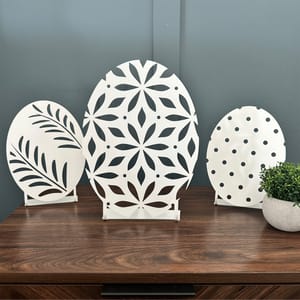 Three white tabletop decorations in the shape of Easter eggs feature intricate cut-out designs: a leaf pattern, a floral pattern, and polka dots, all set on small stands on a wooden table.