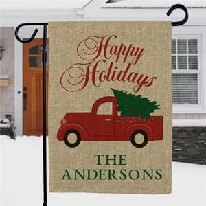 Holiday garden flag with \“Happy Holidays\“ text, featuring a red truck carrying a Christmas tree, with personalized family name \“The Andersons.\“