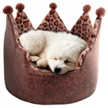 A small white dog is sleeping in a crown-shaped pet bed with leopard print and plush pom-poms.